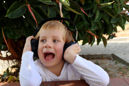 A six-year-old boy with his mouth open listens to music on headphones in the park. Emotional young music lover.の写真素材