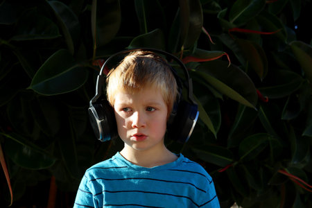 A blond boy listens to music against the background of green plants. A young music lover wearing headphones looks thoughtfully to the side.の写真素材