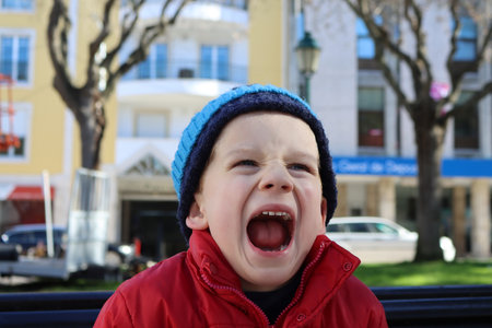 Emotional six year old boy with his mouth wide open. Little boy screaming in a red jacket and blue hat on the streetの写真素材