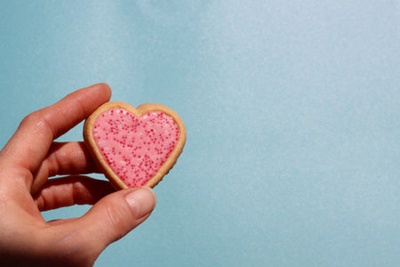 Hand holding a heart shaped cookie on a blue background with copy spaceの写真素材