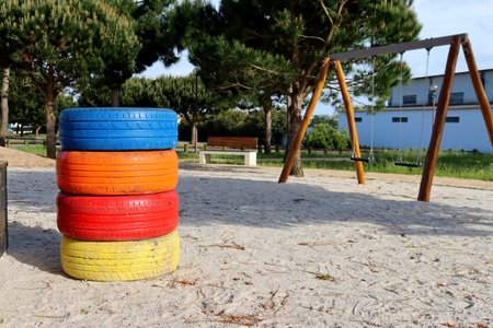 A vertical pile of painted tires in blue, orange, red, and yellow stands on sandy ground with a swing set in the background. Concept of eco-friendly playground design and reuse ofの写真素材