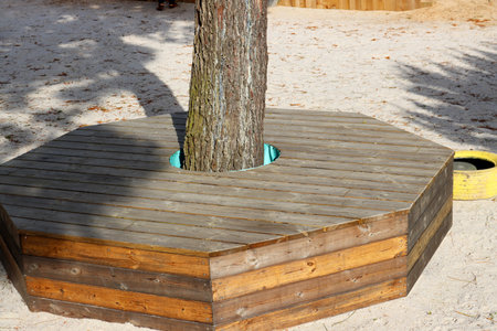 Wooden octagonal bench structure built around a tree trunk on a sandy playground. Serves as both seating and protection for the tree in a recreational outdoor area.の写真素材