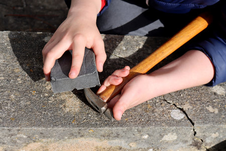 Child holding a hammer and sanding block, working on a stone surface outdoors. Concept of manual work, skill development, and hands-on learning.の写真素材