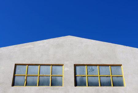 Minimalist facade of industrial building with two yellow-framed windows and bright blue sky, simple...