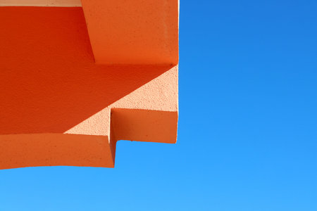 Minimalist geometric architecture with sharp orange angles against a clear blue sky. Strong...