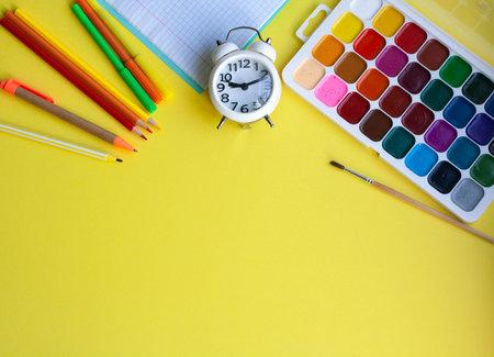 School background with school supplies on yellow pen pencils markers watercolors notebook and alarm...