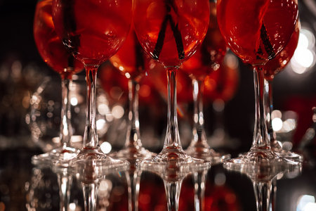 Glasses with an alcoholic cocktail of red color stand in a row on a mirror table. Glasses with...