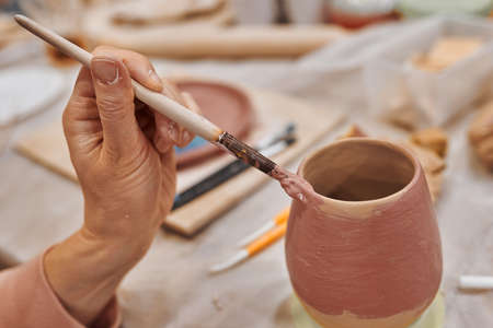 Applying engobe on a ceramic mug with a brush in a pottery workshop. Hands close upの写真素材