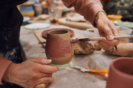 Applying engobe on a ceramic mug with a brush in a pottery workshop. Hands close up