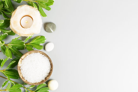 White sea bath salt in a coconut bowl, candle and green leaves on a gray background. Spa and...
