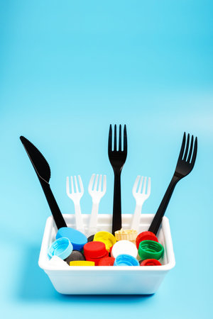 Multi-colored plastic bottle caps and plastic forks in a white plastic box on a blue background. The...