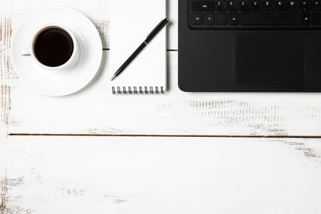 Black laptop, black coffee, notebook, pen on a white wooden table, top view. Home Office. The...