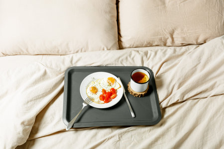 Breakfast in bed. Fried eggs and tea with lemon on a gray tray on a light bedding. Cozy morning at...