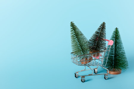 Christmas mini trees in shopping cart on a blue background. Christmas and New Year celebration concept. Holiday shopping and gift shopping concept. Minimalism, copy space.の写真素材