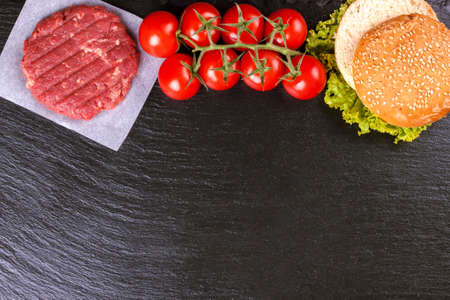 The raw ingredients for the homemade burger. Raw coutlet, tomatoes, cheese, salad, wheat bun on black slate background on the top side with space for text. Top view. Flat lay.の写真素材