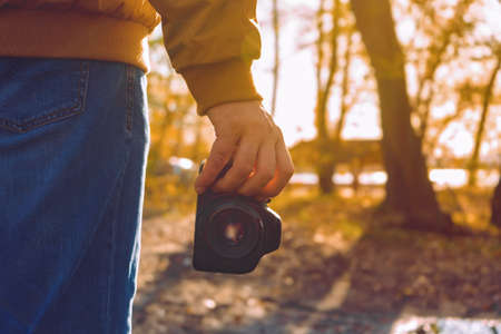 Professional male photographer hold black modern dslr camera in hand with natural fall outside background in sunny day.の写真素材