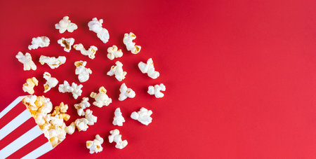 Tasty cheese popcorn falling out of a red striped cardboard bucket, isolated on red background. Scattering of popcorn grains. Movies, cinema, fast food and entertainment concept. Top view, flat layの写真素材