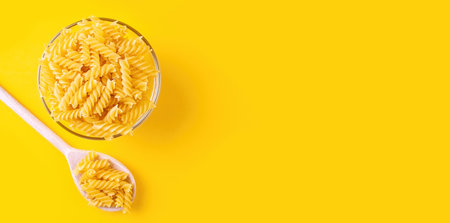 Fusilli spaghetti pattern, food background. Bowl and wooden spoon with pasta on yellow background, banner, header, template with copy space. Raw pasta, ingredient for cook, traditional italian cuisineの写真素材