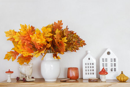 Bright orange and yellow autumn leaves in white vase on wooden table top. Nearby decorative houses and candle on white plaster wall background. Fall decor for Halloween, Thanksgiving day holidays.の写真素材