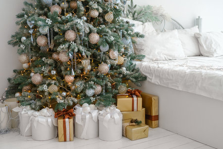 Holiday morning in bright, decorated room with Christmas tree with ornaments and lights and wrapped giftboxes gathered by bed with white bedding, creating a festive atmosphere. Background, wallpaperの写真素材