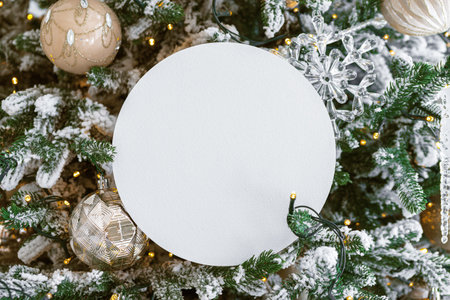 White blank paper circle on festive decorated Christmas tree background. Mockup, template, copy space for text. Colorful holiday decorations, red berries, green leaves, and twinkling lights.の写真素材