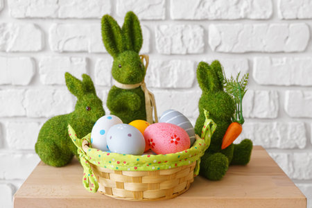 Trhee decorative green moss bunny figures with colorful Easter eggs in wicker basket on wooden table on white brick wall background. Easter holiday festive atmosphere, wallpaper, backdrop.の写真素材