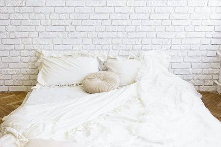 Cozy white bedding, bed with pillows on white brick wall background. Minimalist bed with soft white blanket, plush round pillow and rectangular cushions creating a warm and inviting atmosphere in the room.の写真素材