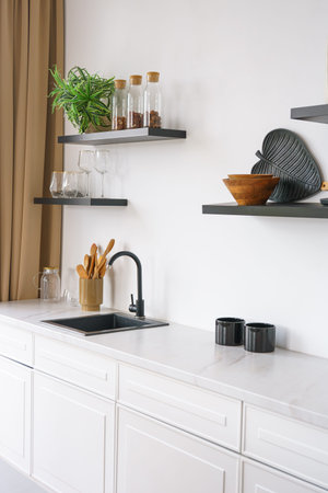 White kitchen with countertop and black water tap on white wall background with wooden shelves. Shelves with utensils, kitchenware and decor. Design interior, minimalist styleの写真素材