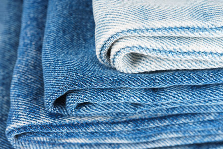 Pile of folded textiles, various shades of blue jeans fabric, denim cloth, are stacked on top of each other in store setting, showing different textures and patterns. Textured background, backdropの写真素材