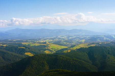 Atmospheric view of multiple mountain ridges covered with green coniferous forest and valleys, fading into distance under cloudy sky. Summer haze and soft sunlight create peaceful and majestic scene.の写真素材