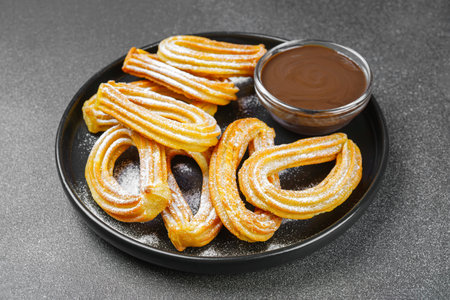 Traditional spanish dessert churros, fried dough pastry dusted with powdered sugar and chocolate sause on black background. Street food, sweet snack, homemade dessert.の写真素材
