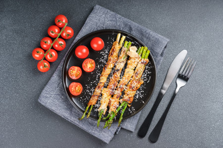 Delicious crispy bacon wrapped asparagus served with fresh cherry tomatoes on black plate and gray background. Perfect food photography for culinary blogs, menus, or healthy meal promotions.の写真素材