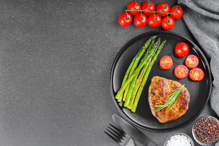 Tasty grilled beef steak served with green asparagus and cherry tomatoes on black plate on gray textured backgound. Top view, copy space. Perfect for food, diet, or culinary design concepts.の写真素材