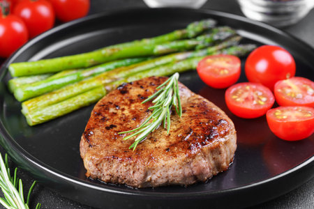 Close up, macro of tasty grilled beef steak served with green asparagus and cherry tomatoes on black plate, with cutlery. Perfect for food menu, diet, or culinary design concepts.の写真素材