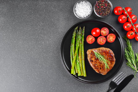 Tasty grilled beef steak served with green asparagus and cherry tomatoes on black plate on gray textured backgound. Top view, copy space. Perfect for food, diet, or culinary design concepts.の写真素材