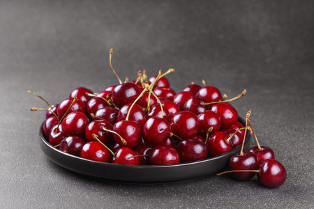 Close up of black plate filled with ripe red sweet cherry placed on dark gray textured background. Delicious food, juicy summer fruit and healthy eating.の写真素材