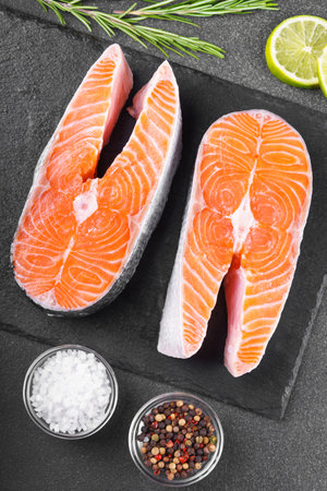 Two fresh raw salmon or trout steaks on black stone surface with food ingredients. Tomatoes, lime, rosemary, salt, and pepper. Healthy eating, seafood, cooking, recipe and menu concepts.の写真素材