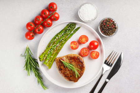 Tasty grilled beef steak served with green asparagus and cherry tomatoes on white plate, with cutlery and spices. Perfect for food menu, diet, or culinary design concepts. Top view, flat layの写真素材