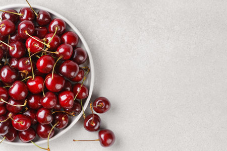 Close up of black plate filled with ripe red sweet cherry placed on light gray textured background....