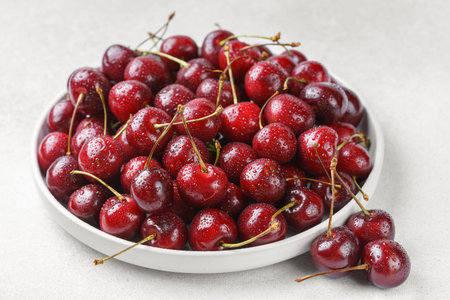 Close up of white plate filled with ripe red sweet cherry covered in fresh water drops, placed on...