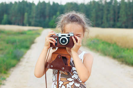 Happy beautiful little girl in retro outfit is taking pictures with old film camera.の写真素材
