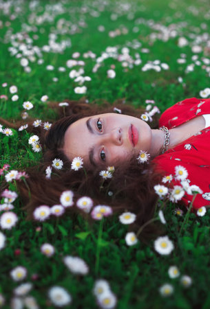 Young beautiful girl in red dress is resting on fresh spring grass with little white flowers.