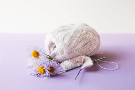 A soft pink ball of thread with knitting needles and purple erigeron flowers on a purple background