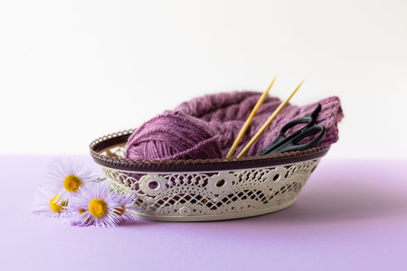 A purple ball of thread and a knitted hat with knitting needles and scissors in a basket on a purple...