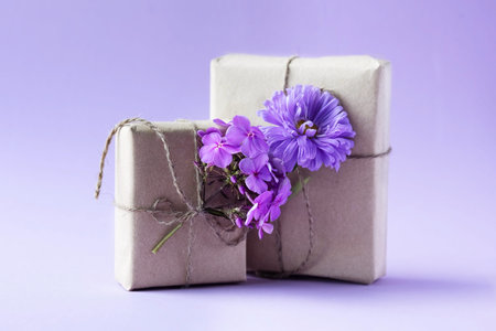 Two gift boxes in gray craft paper decorated with lilac aster flowers on a purple background for the...