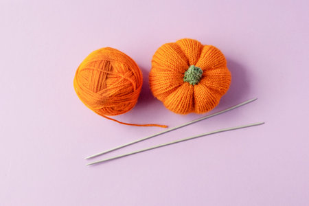 Knitted orange pumpkin and an orange ball of thread with knitting needles on a purple background,...