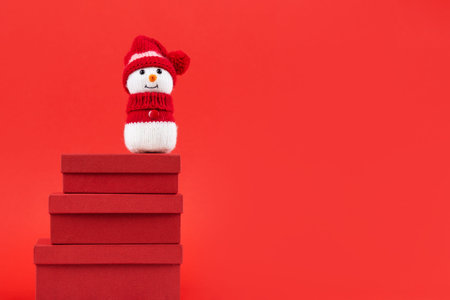 Knitted snowman in a red hat and sweater stands on three red gift boxes on a red background. Merry Christmas and New Year 2023 greeting cardの写真素材