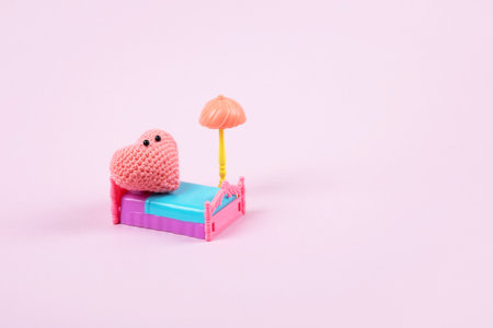 Concept of Valentines Day. A composition of a knitted pink heart that sleeps on a pink toy bed with a blue blanket under a toy lamp on a lilac background. Symbol of love, family, loyaltyの写真素材