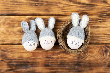 Three Easter eggs with funny faces and gray crocheted hats with bunny ears in a decorative nest of...