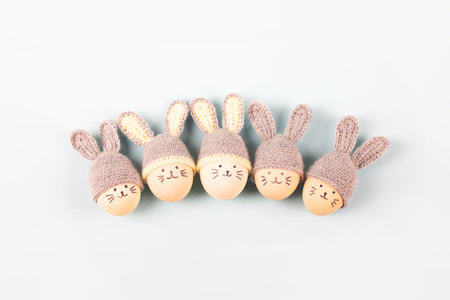 Five Easter eggs with funny faces and gray crocheted hats with bunny ears on a gray background....
